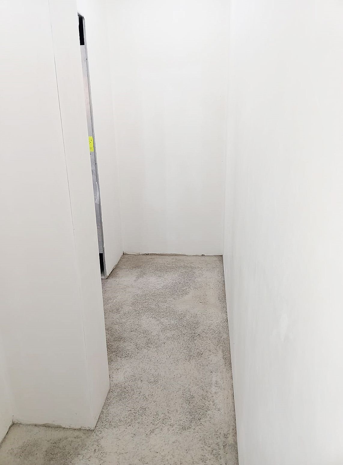 Corridoio stretto con pavimento in cemento grezzo e pareti bianche, che conduce a una porta.