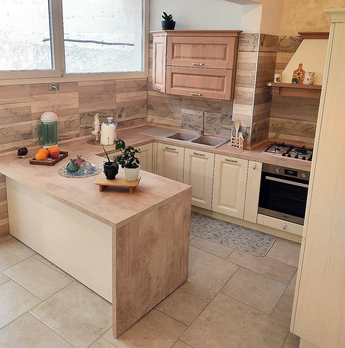 Cucina con mobili beige, paraschizzi piastrellato, isola con piano di lavoro e finestra.