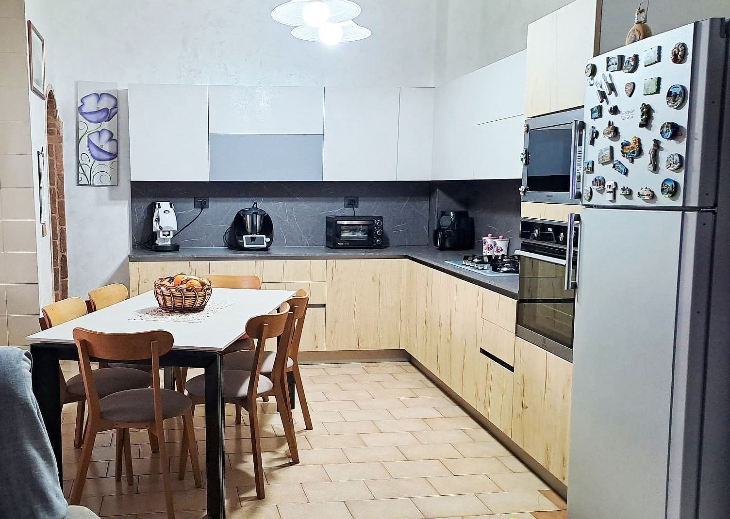 Cucina con mobili bianchi e in legno, elettrodomestici, tavolo da pranzo con sedie e quadri alle pareti.