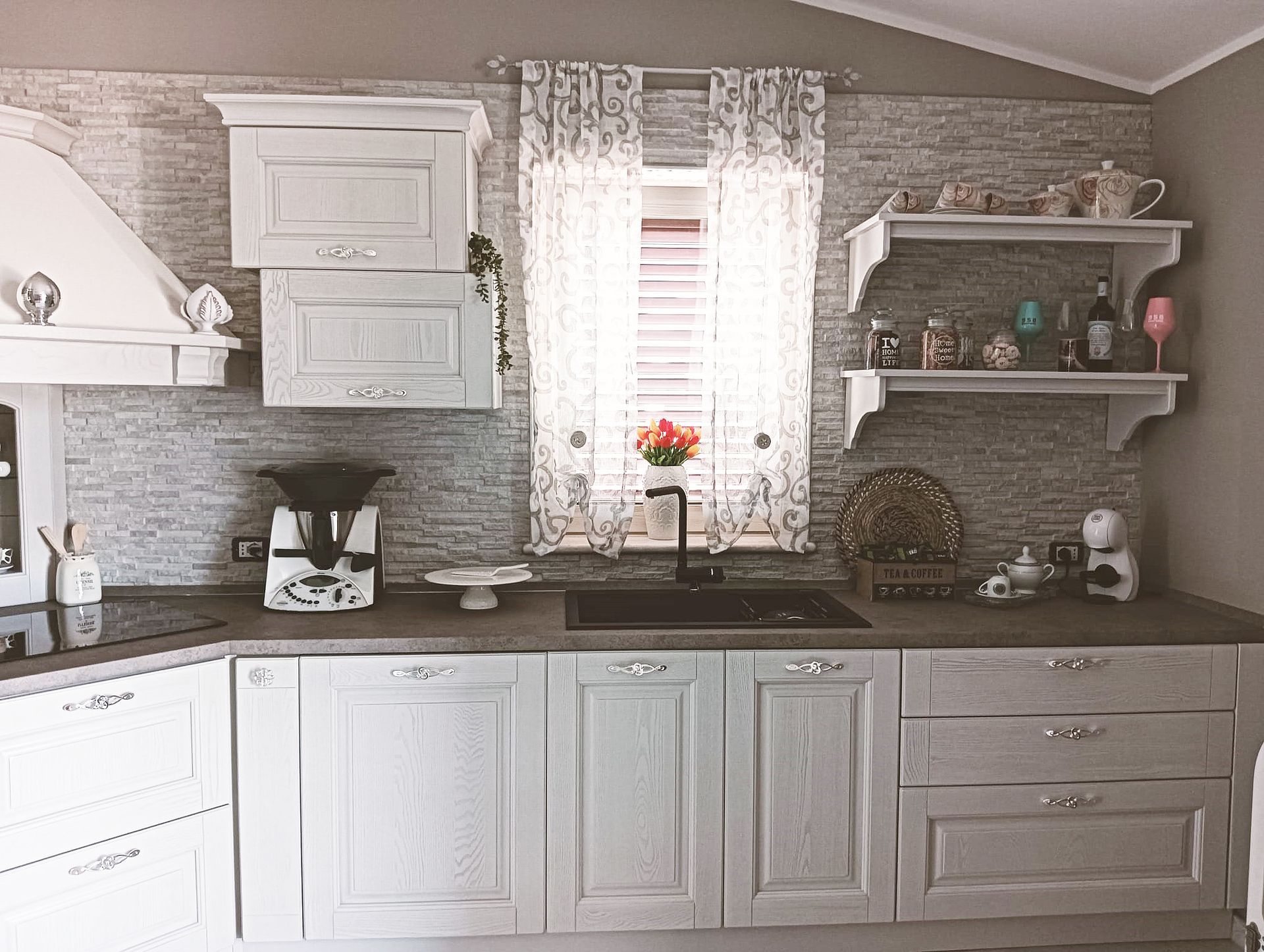 Cucina grigio chiaro con paraschizzi in pietra, mobili e scaffali a giorno con oggetti decorativi.