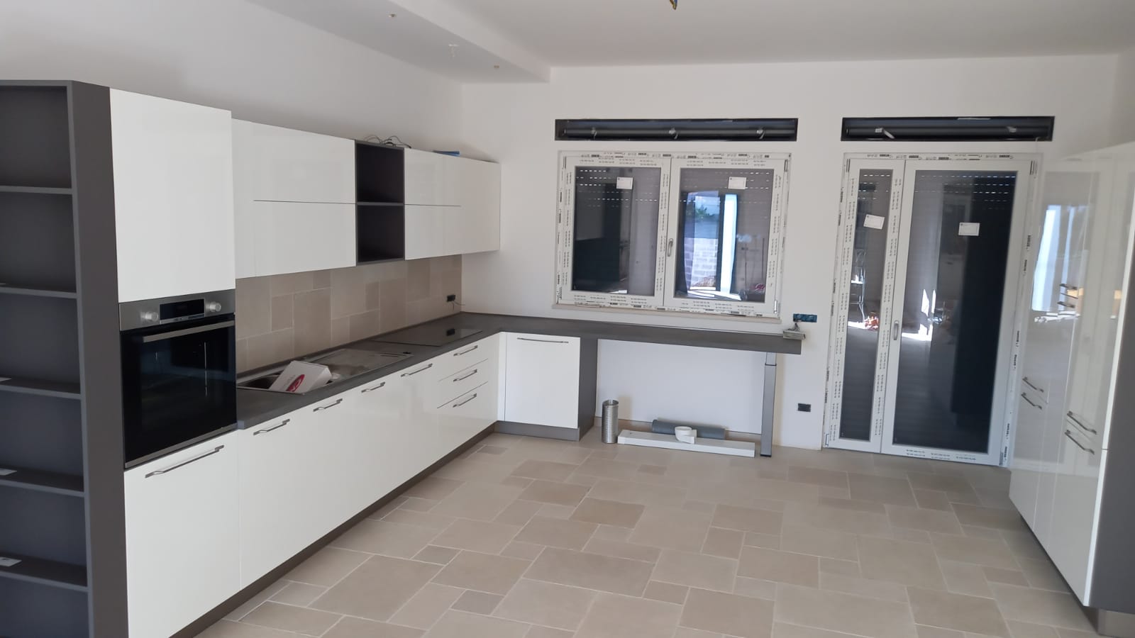Moderna cucina a L con mobili bianchi, dettagli grigi e pavimento piastrellato, vicino alle finestre.