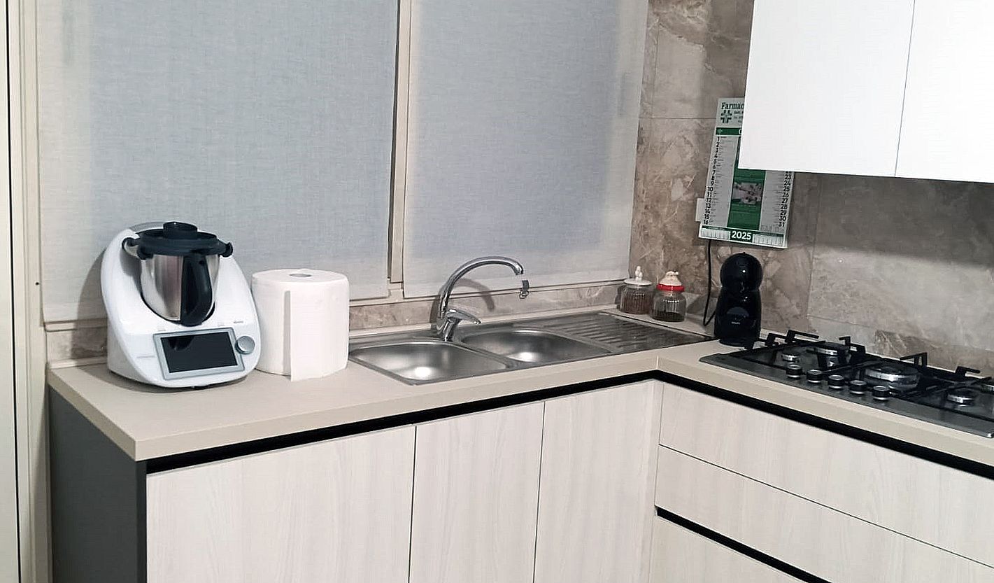 Cucina con lavello, fornelli e Thermomix su un piano di lavoro di colore chiaro; finestra sullo sfondo.