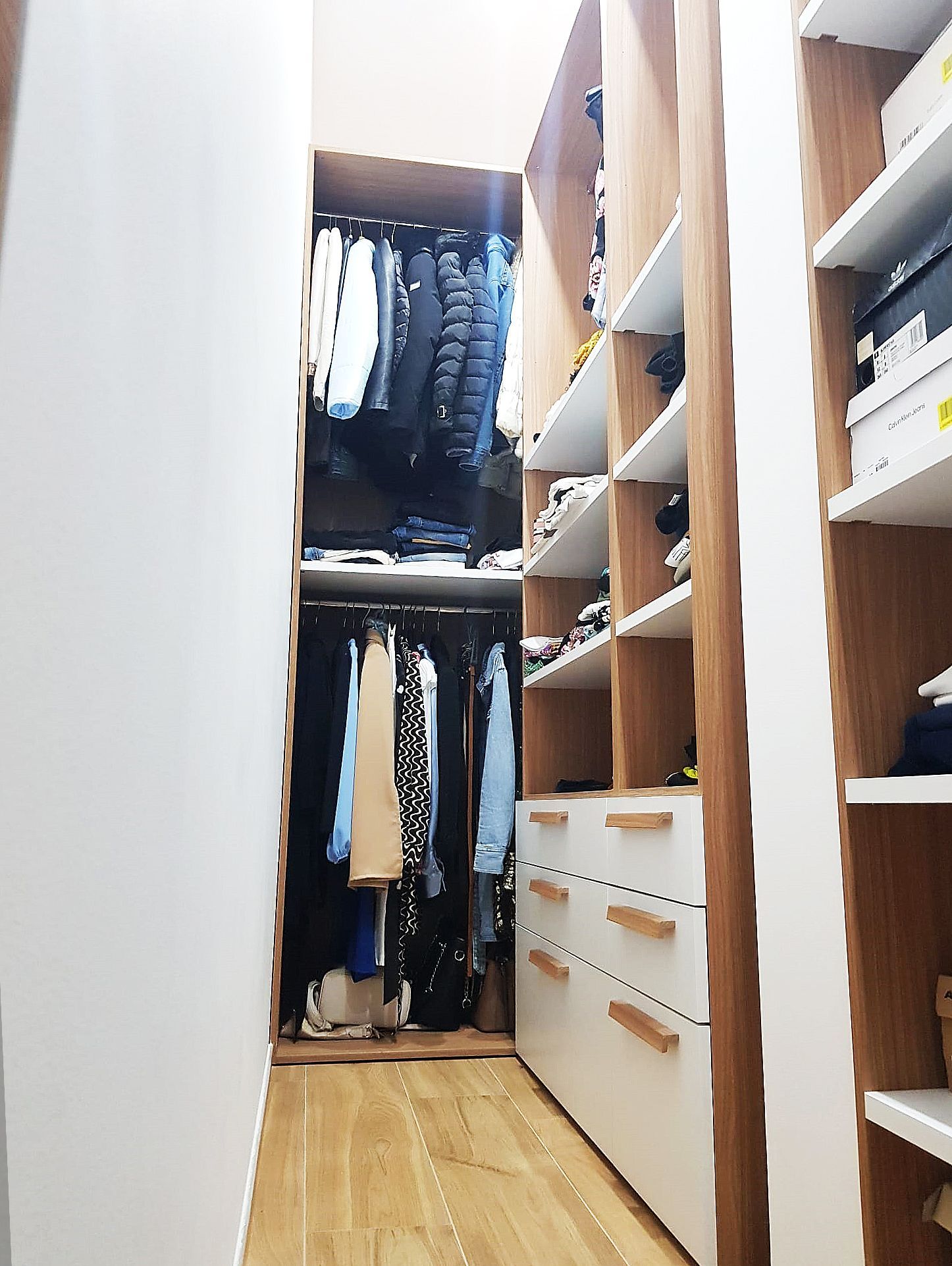 Cabina armadio con abiti appesi, ripiani per scarpe e cassetti; design in legno e bianco.