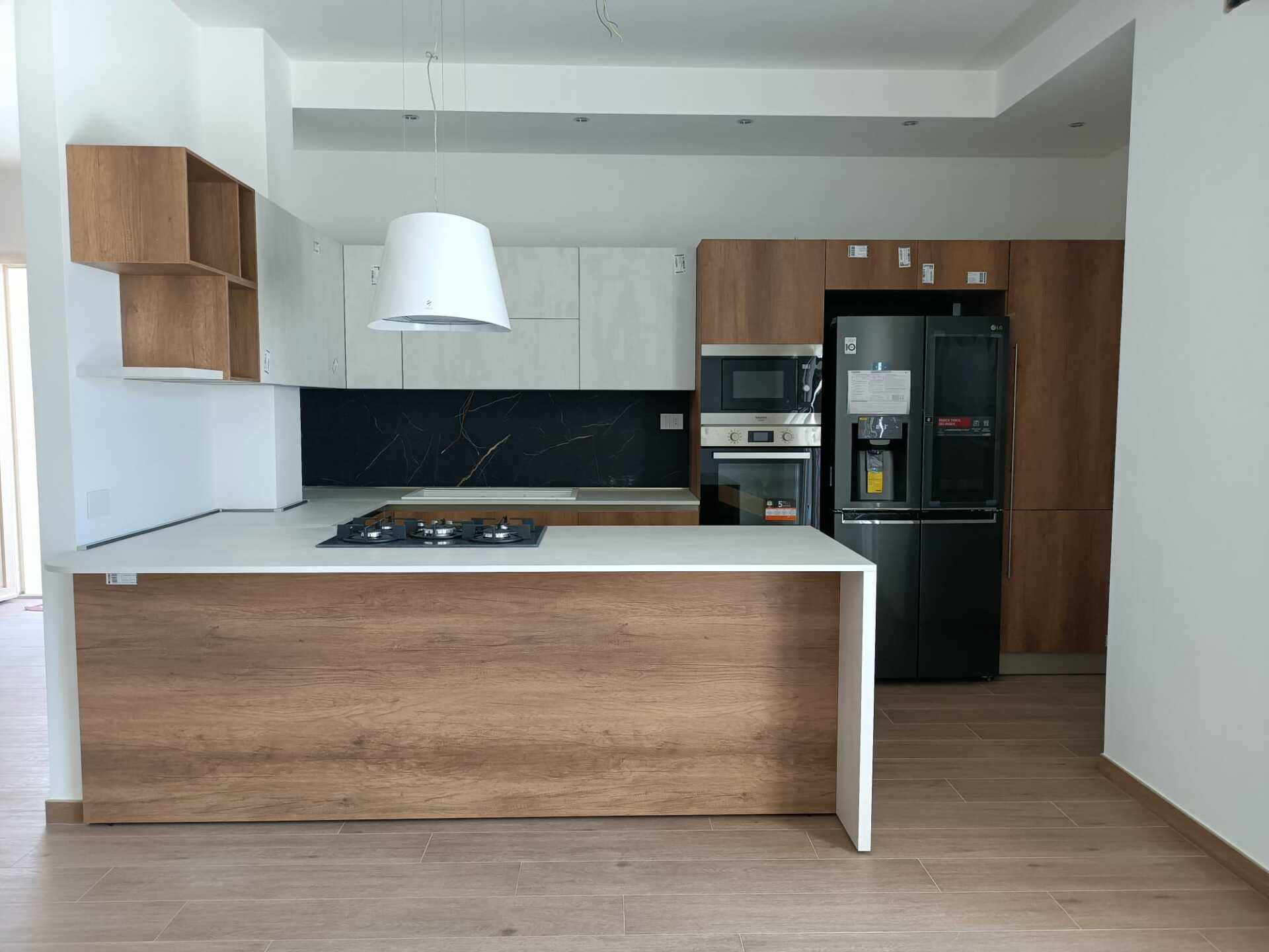 Cucina moderna con mobili in legno e bianchi, isola con piano cottura, elettrodomestici neri e piano di lavoro bianco.
