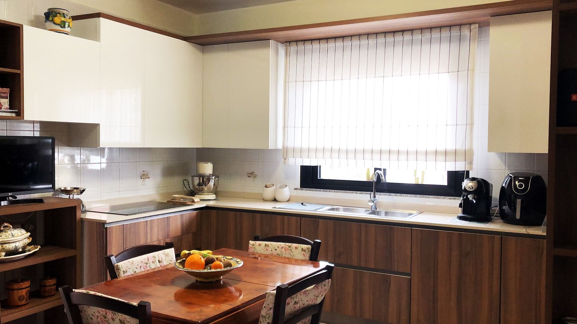 Cucina con mobili bianchi e in legno, lavello in acciaio inox e tavolo da pranzo con frutta.