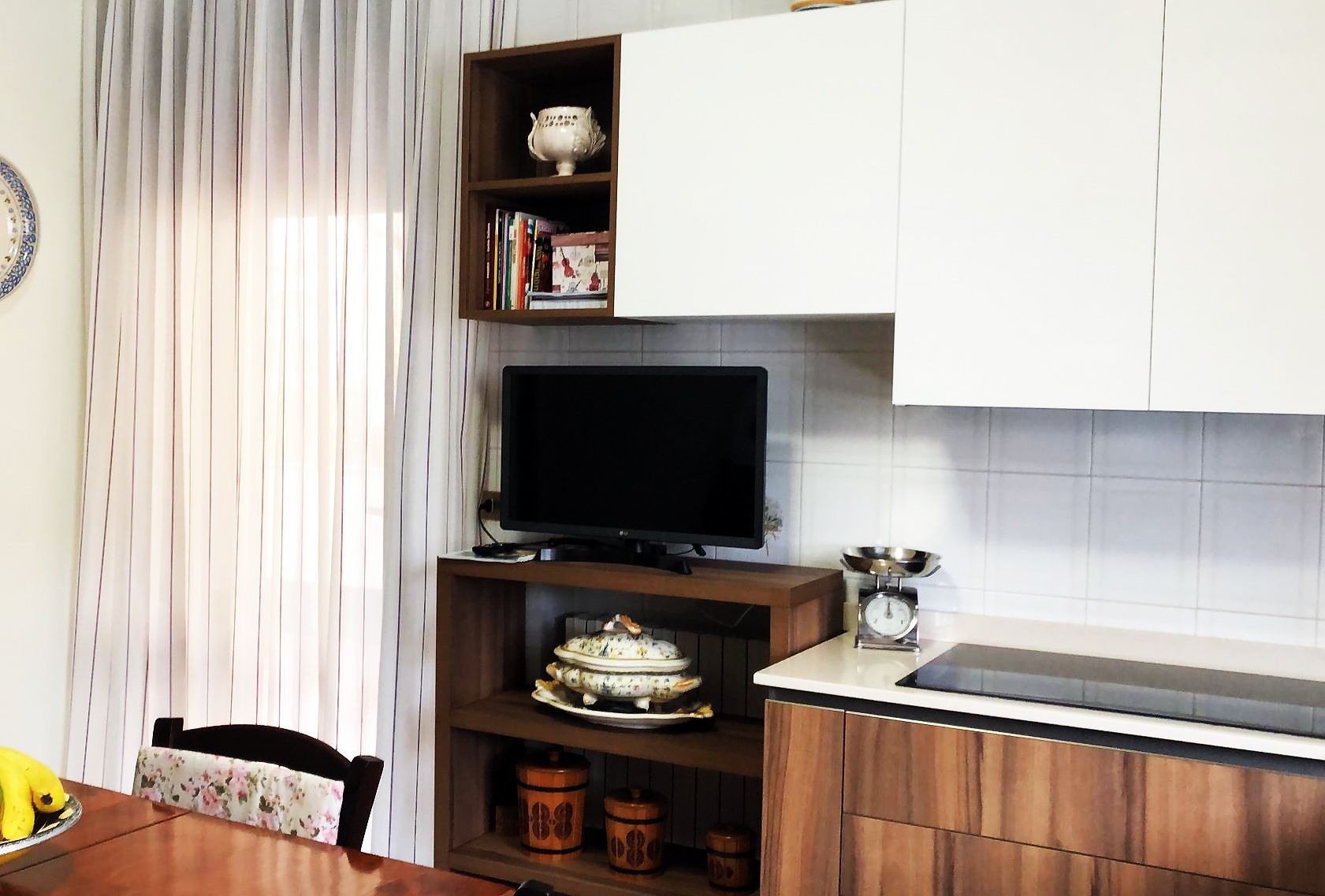 Zona cucina con mobili bianchi, ripiani in legno scuro, TV, finestra con tende trasparenti e tavolo da pranzo.