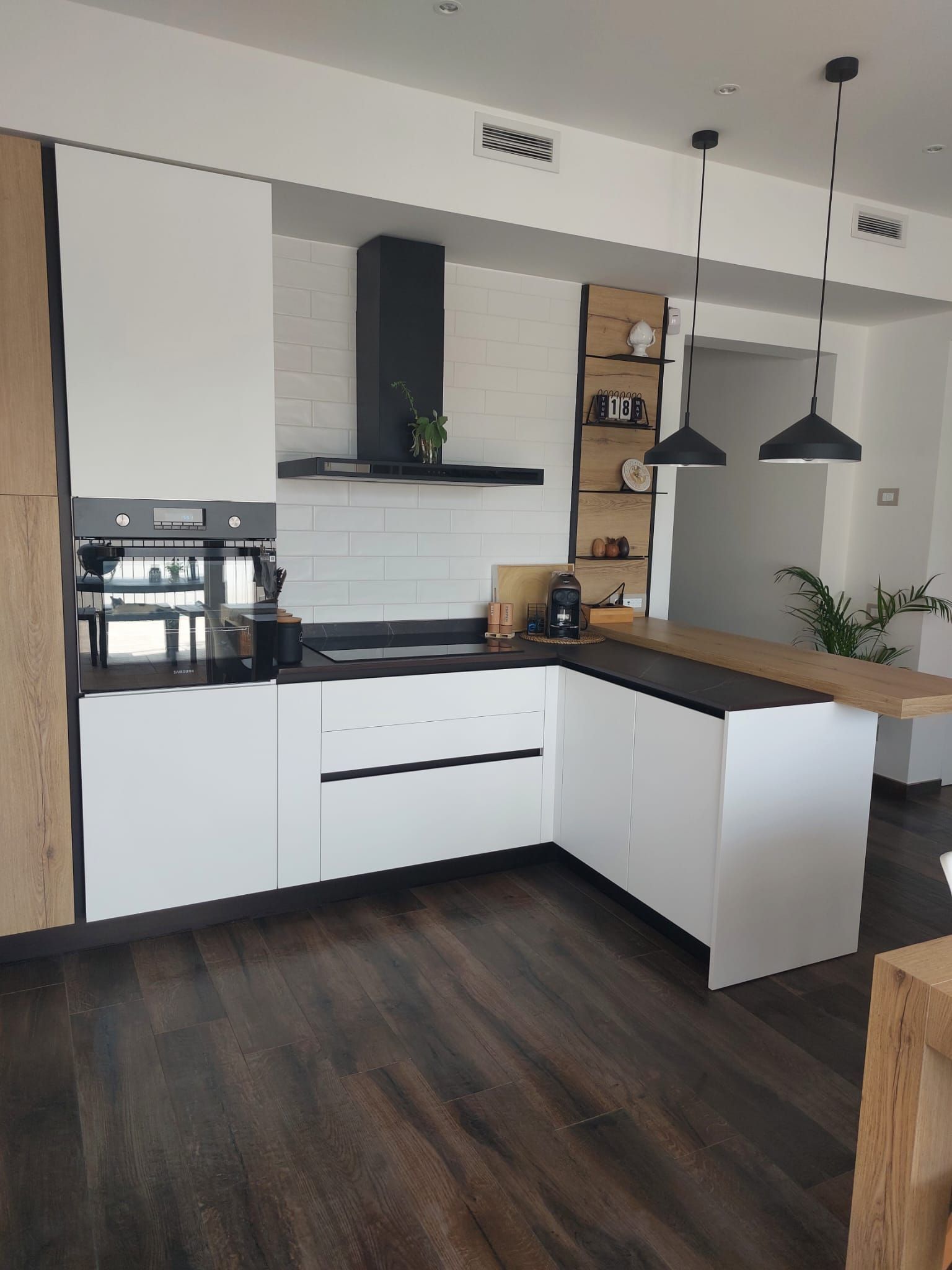 Moderna cucina bianca con ripiani neri, dettagli in legno e lampade a sospensione.