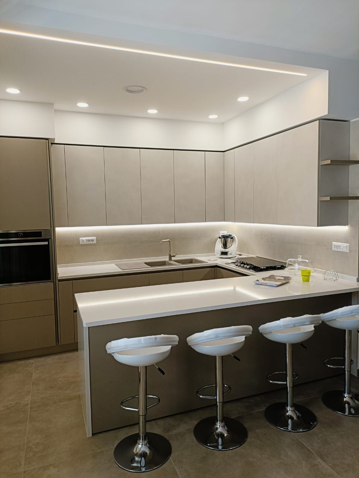 Cucina moderna con mobili beige chiaro, ripiani bianchi e sgabelli da bar. L'illuminazione a LED accentua lo spazio.
