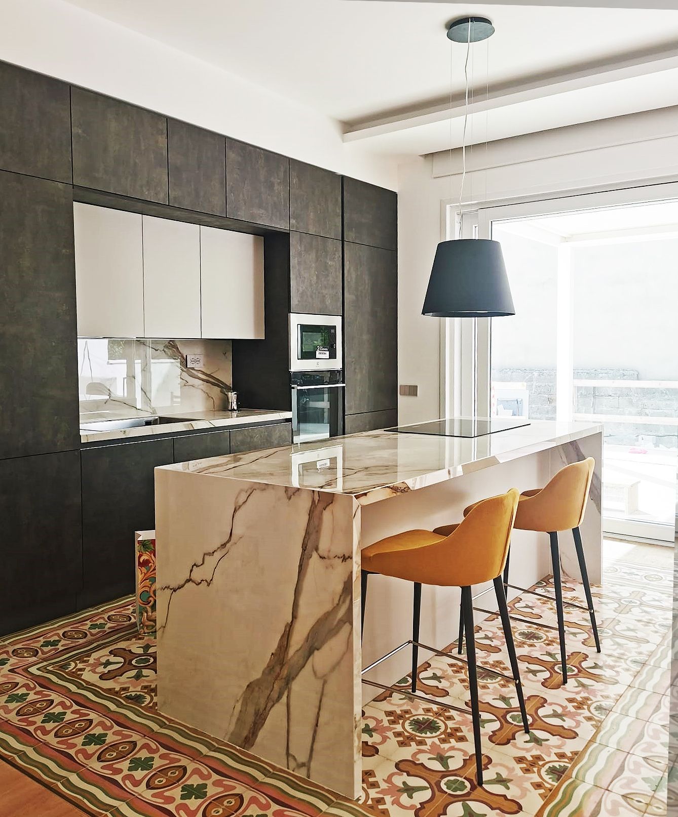 Cucina moderna con isola in marmo, mobili scuri, due sgabelli gialli e tappeto a fantasia.