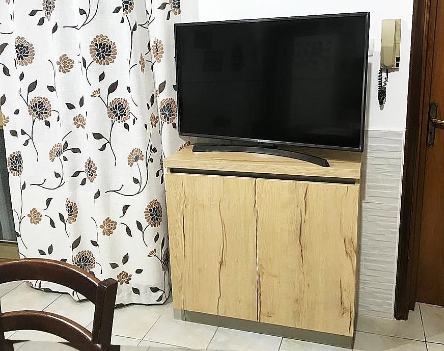 TV sopra un mobile in legno chiaro, con tenda floreale e parete con telefono.