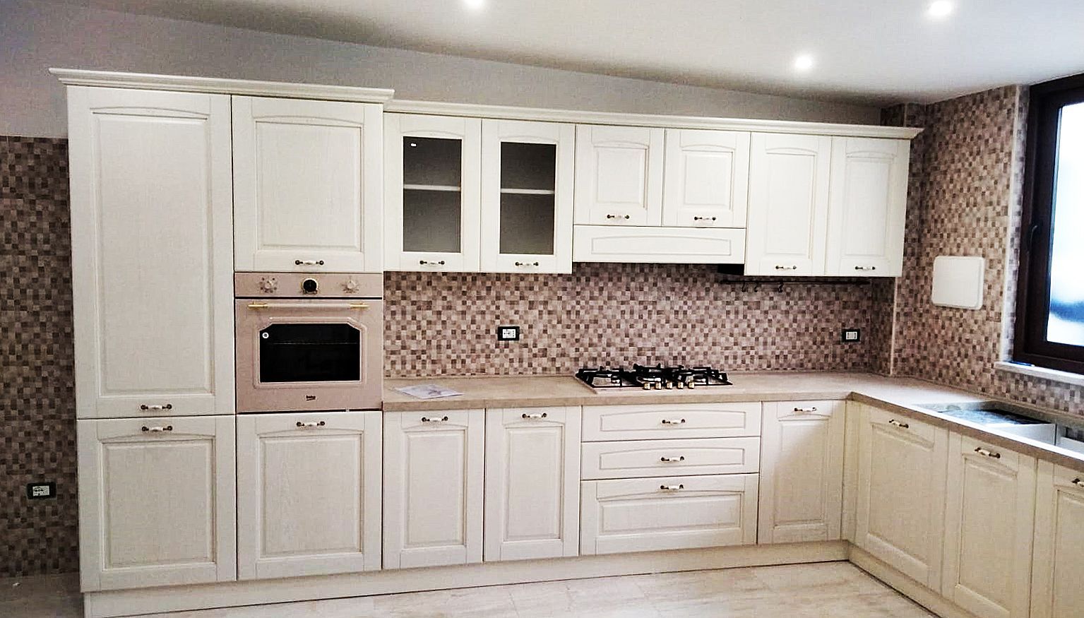 Mobili da cucina color crema con forno, piano cottura e paraschizzi in mosaico. Sono visibili il piano di lavoro angolare e una finestra.