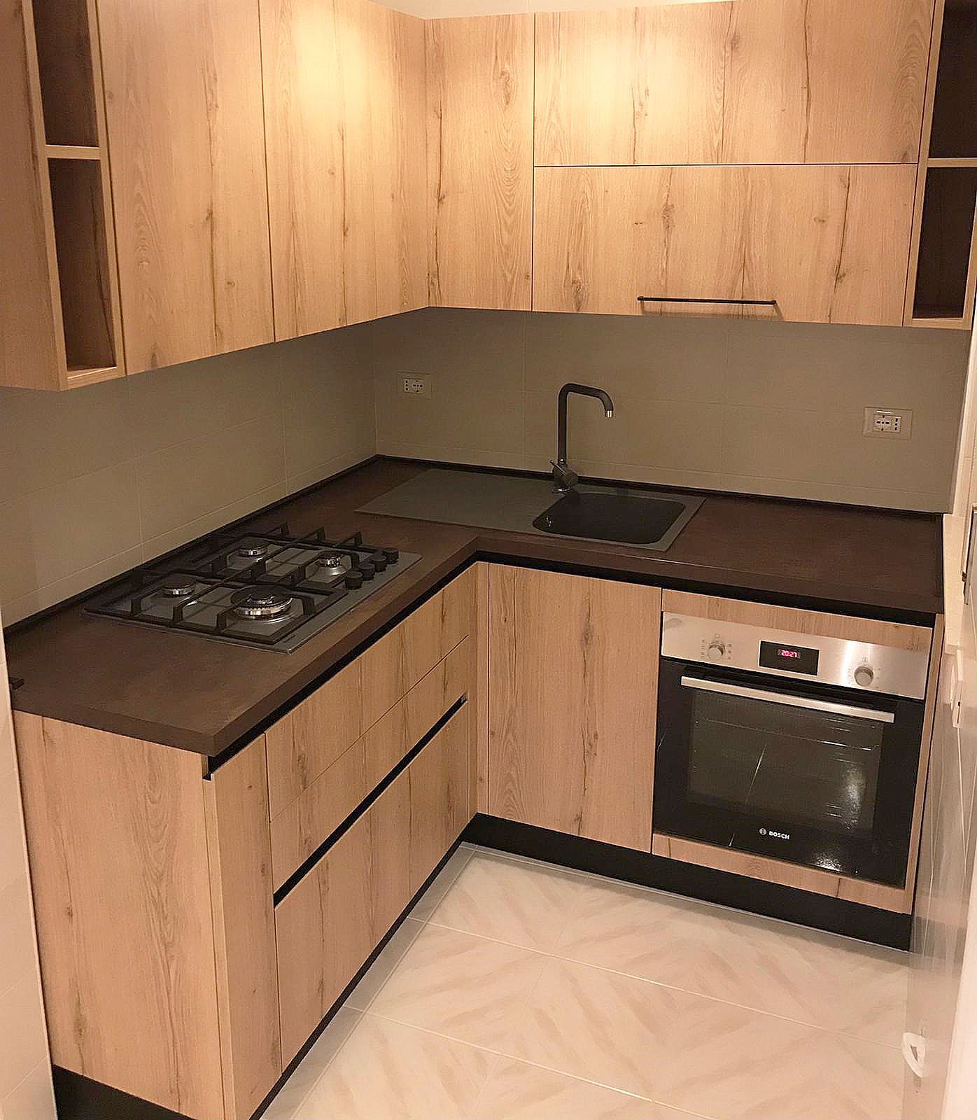 Cucina a L con mobili in legno chiaro, ripiani scuri, lavello, piano cottura e forno.