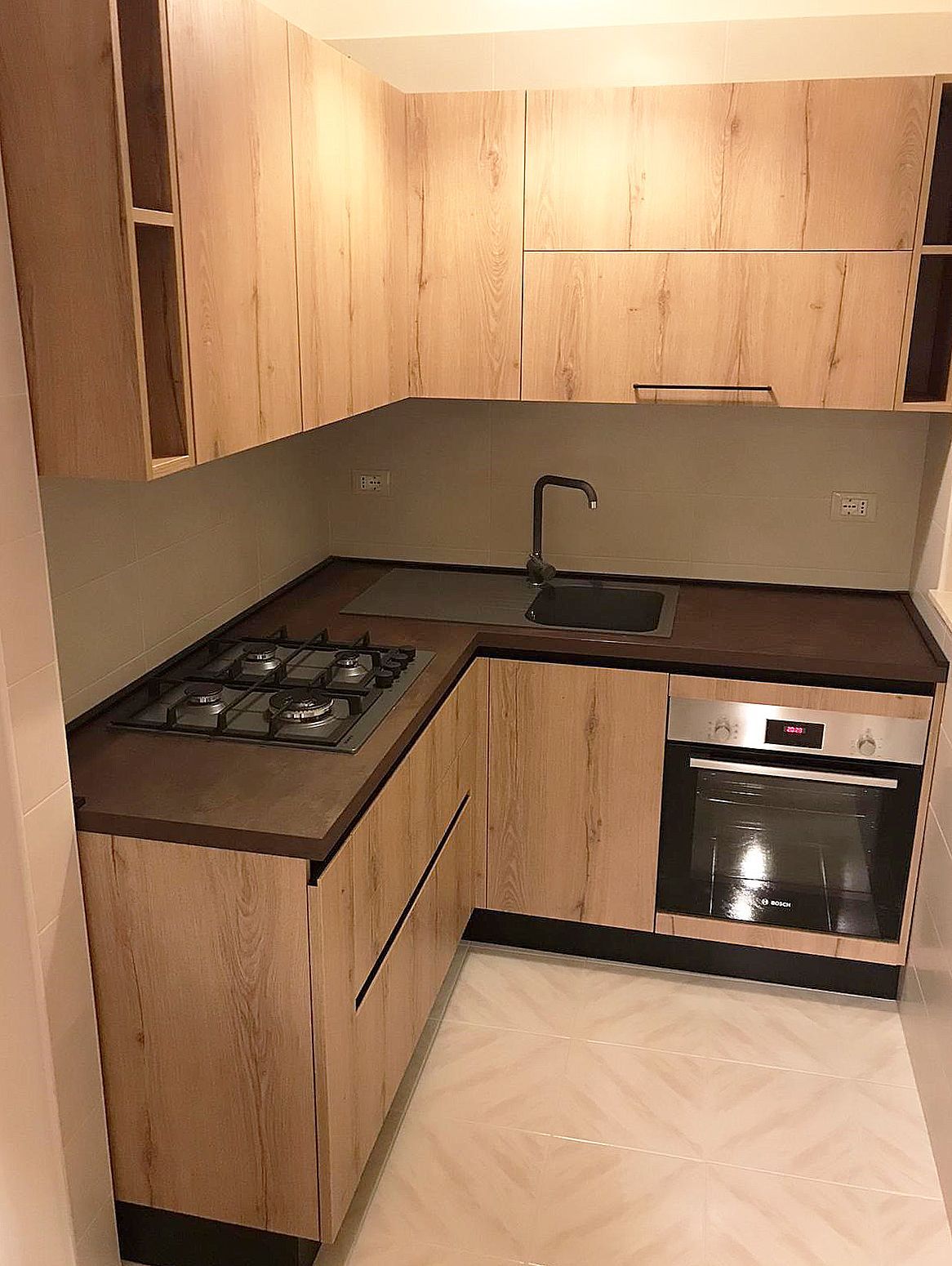 Cucina a L con mobili in legno chiaro, ripiani scuri, fornelli, lavello e forno.