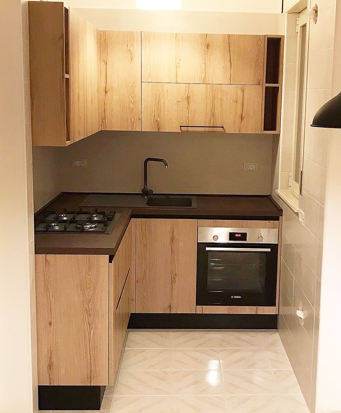 Piccola cucina con mobili in legno chiaro, ripiani scuri e forno nero.