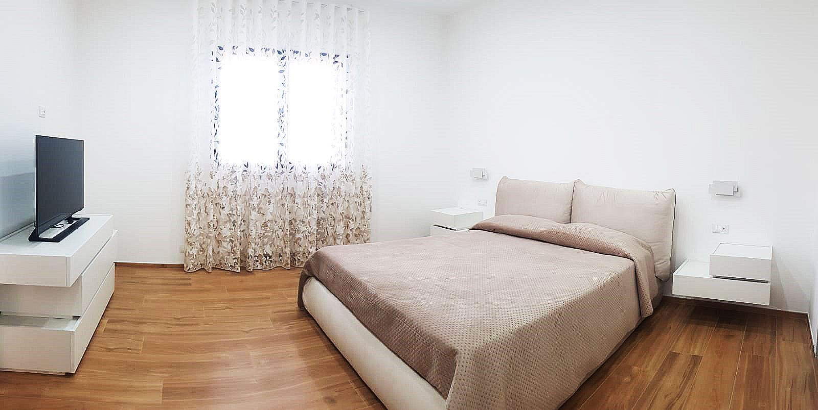 Camera da letto con letto, televisione e mobili bianchi. Pavimento in legno chiaro. Finestra con tende a fantasia.