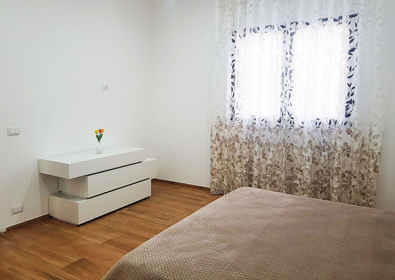 Interno della camera da letto con cassettiera bianca, letto e finestra con tende floreali. Pavimento in legno.