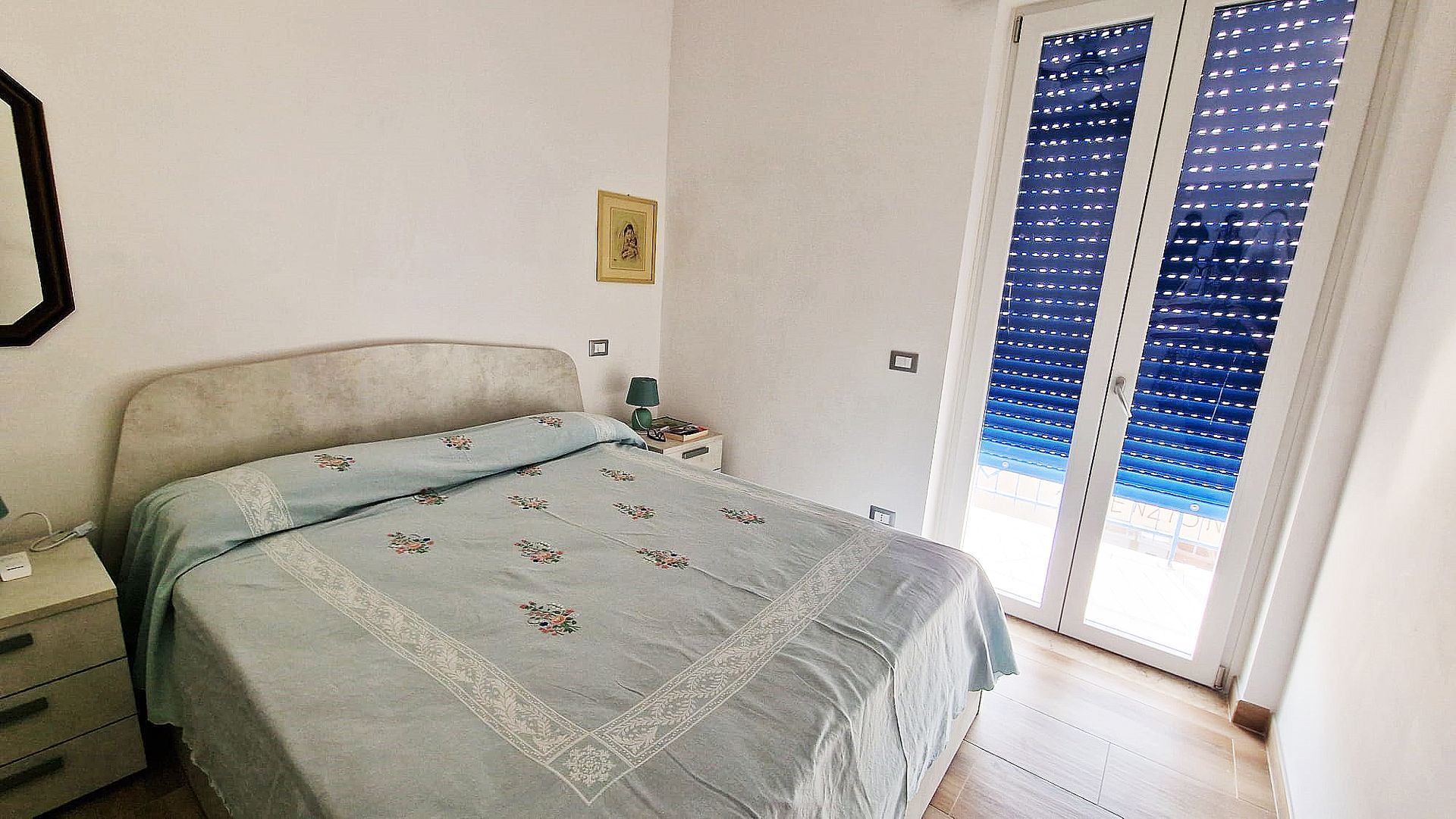 Camera da letto con letto, comodino, specchio e una porta con persiane blu.