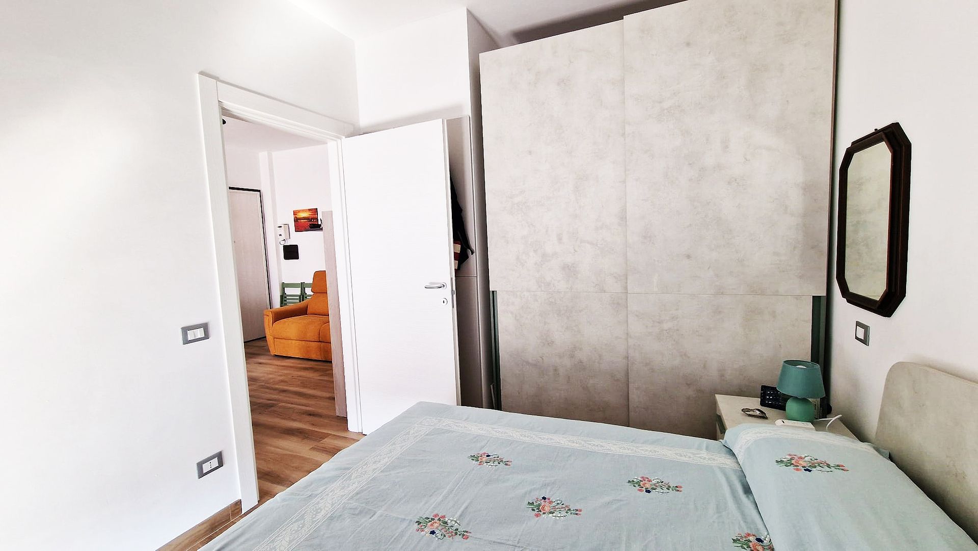 Camera da letto con letto, armadio, porta aperta su un'altra stanza. Copriletto verde acqua, pareti bianche, pavimenti in legno.