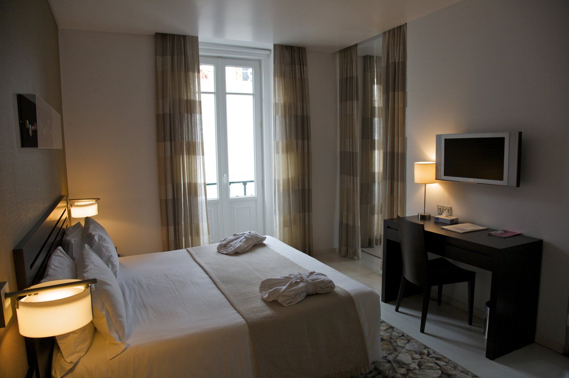 Camera d'albergo con letto, scrivania, TV e finestra con tende trasparenti.