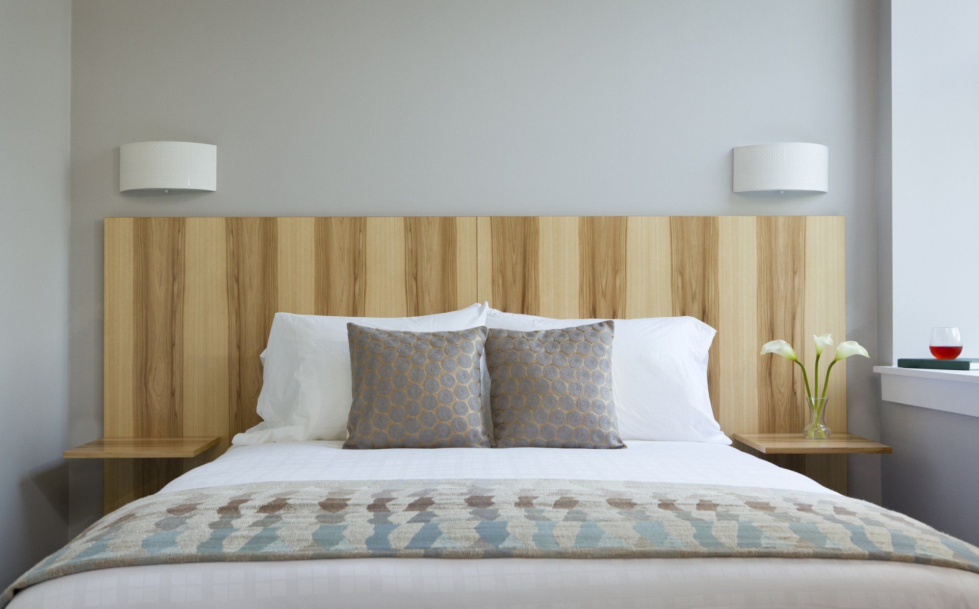 Camera da letto con testiera in legno, due lampade, biancheria da letto bianca, due cuscini grigi e un vaso di fiori bianchi.