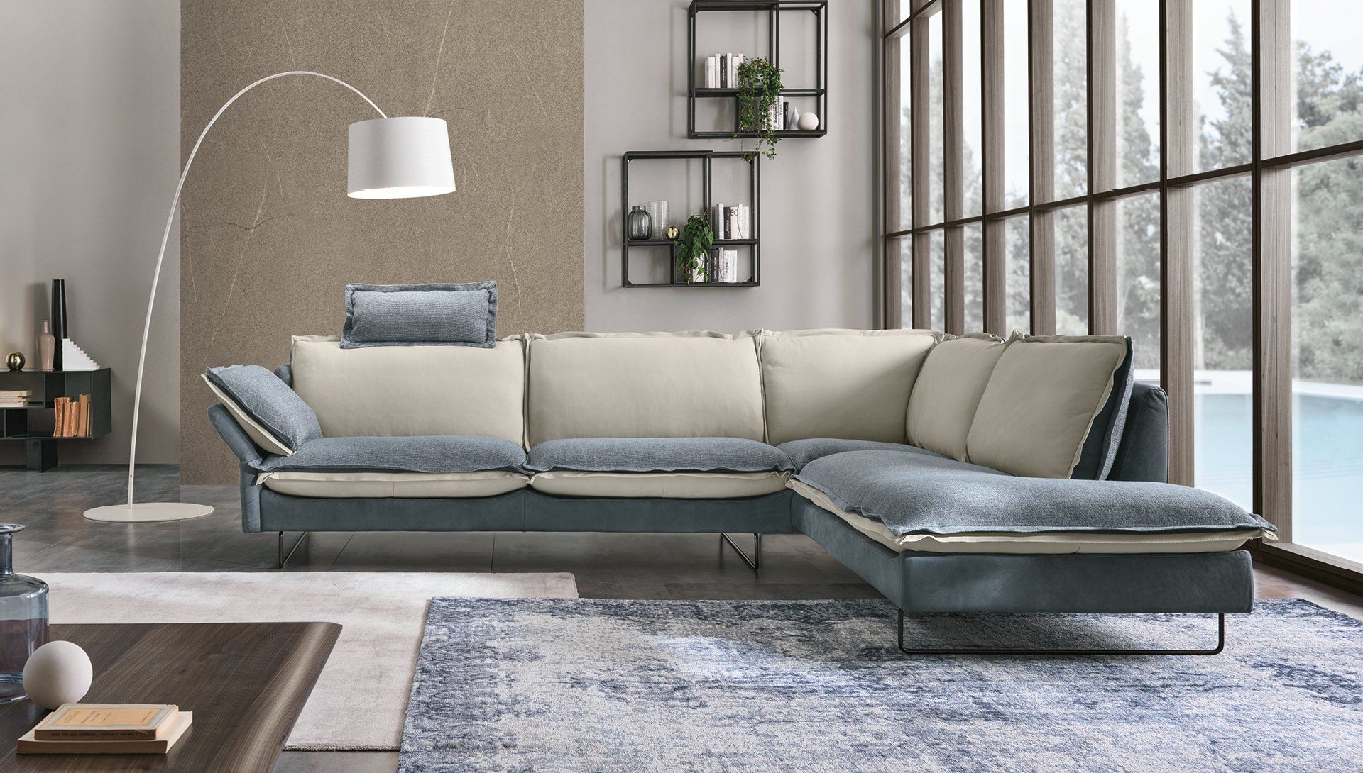 Divano componibile blu e beige in un soggiorno, con lampada da terra e ampia finestra con vista sulla piscina.