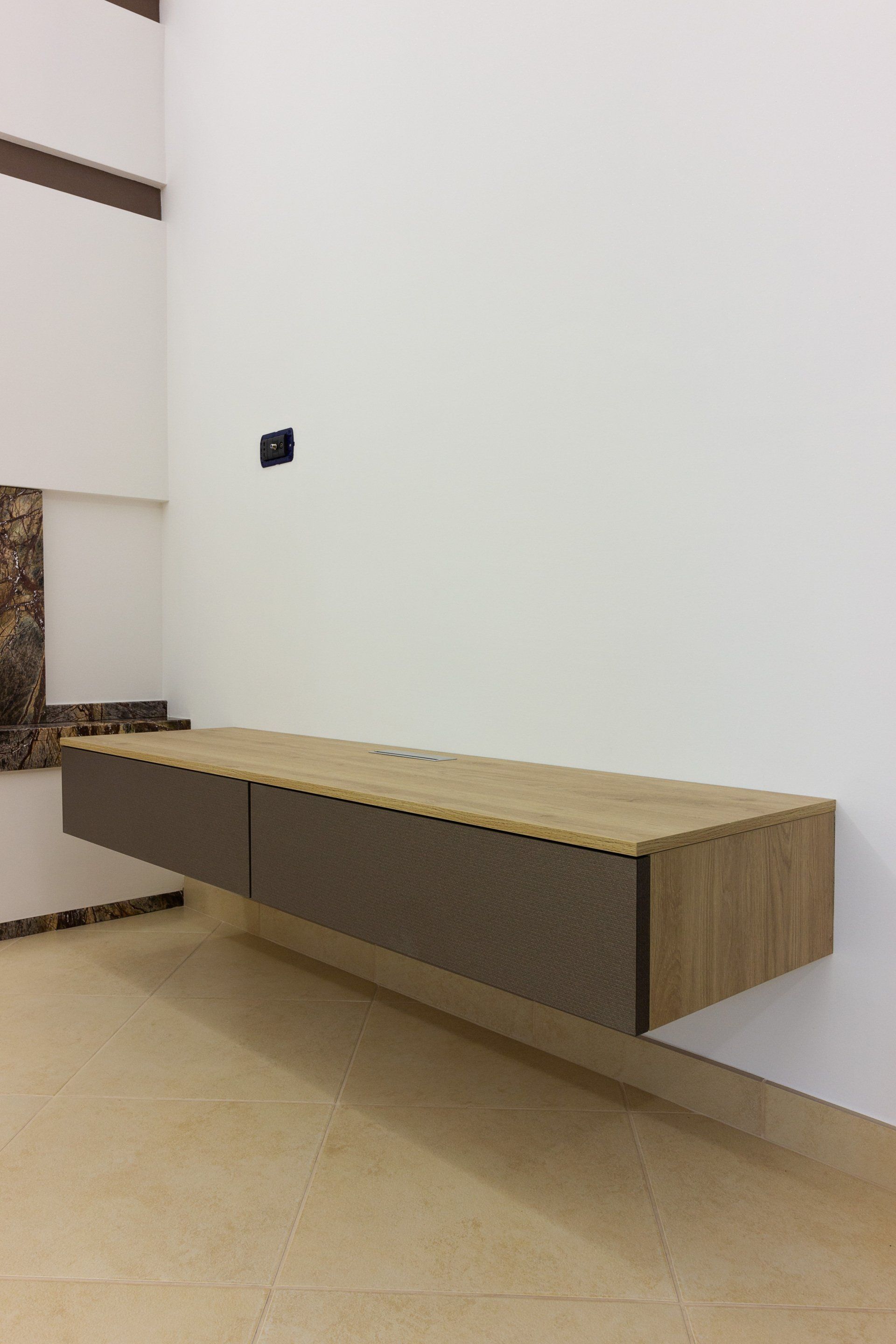 Mobiletto in legno sospeso su una parete bianca, con piano marrone chiaro e cassetti marrone scuro. Pavimento piastrellato beige.