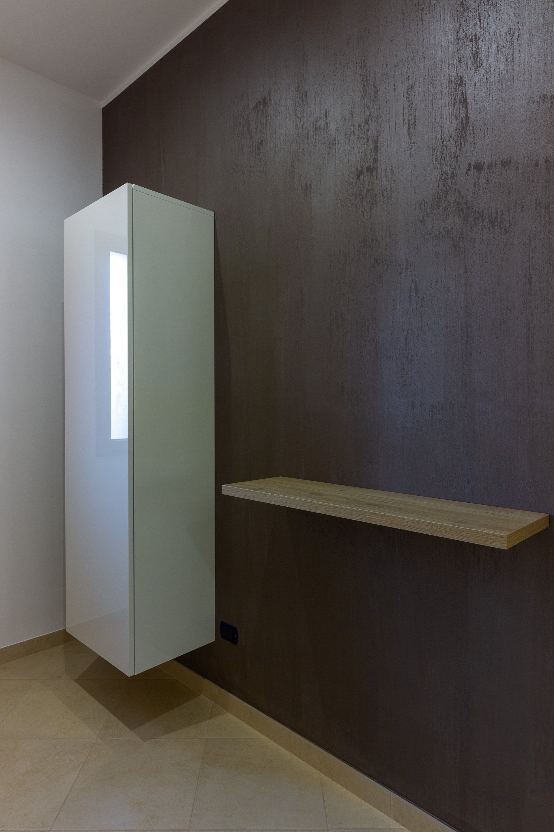 Mobiletto bianco sospeso e mensola in legno su una parete con texture marrone, pavimento color cuoio.