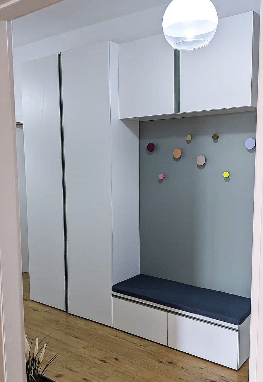 Mobiletto d'ingresso integrato con armadietti bianchi, una panca e ganci colorati su una parete grigia.