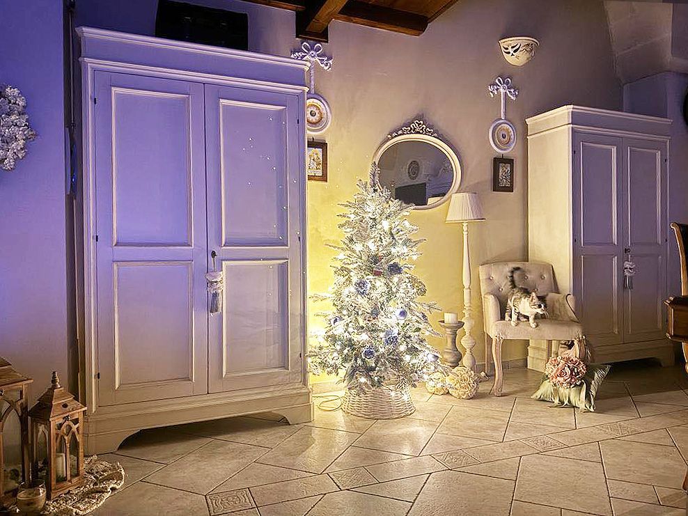Armadi dipinti di bianco fiancheggiano un albero di Natale decorato in una stanza caldamente illuminata, con uno specchio e una piccola sedia.