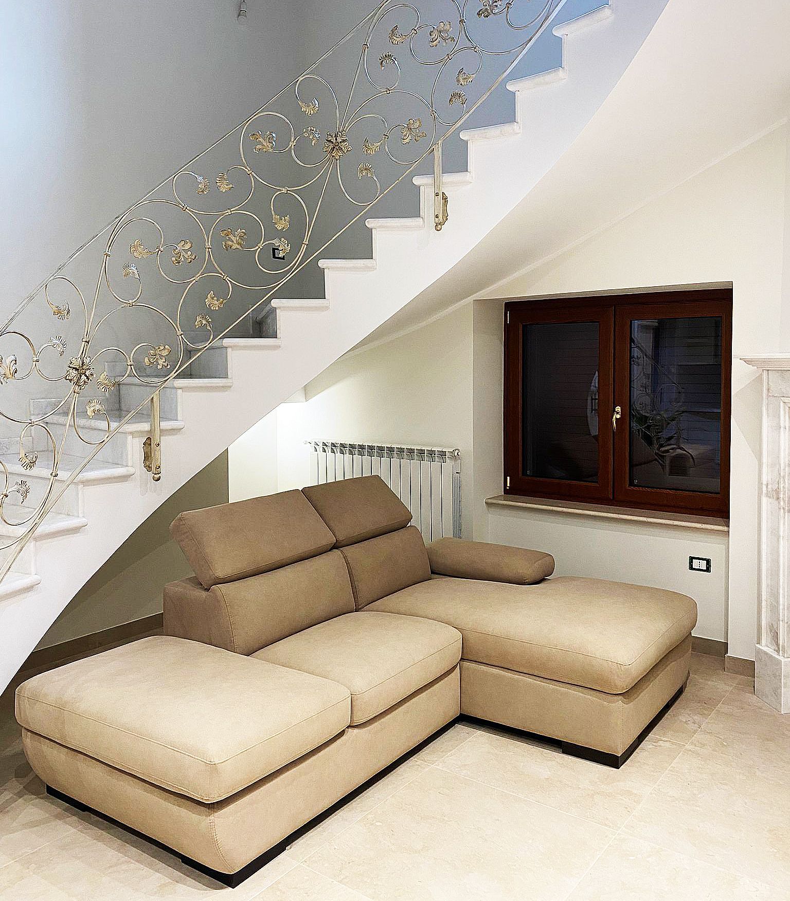 Divano componibile beige sotto una scala con ringhiera decorata in oro e bianco, finestra sulla destra.