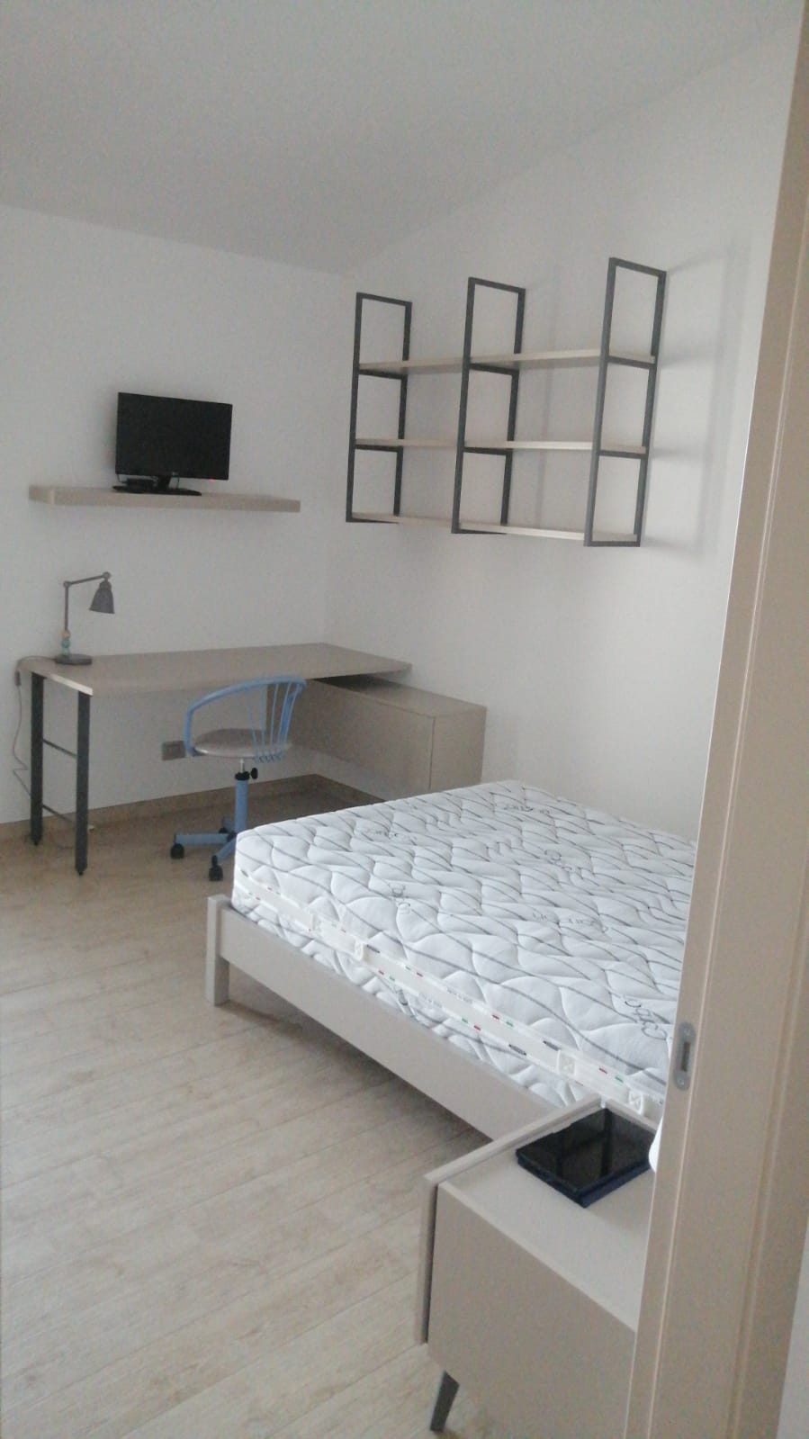 Camera da letto con letto, scrivania, scaffali e televisione. Colori neutri e pavimento chiaro.