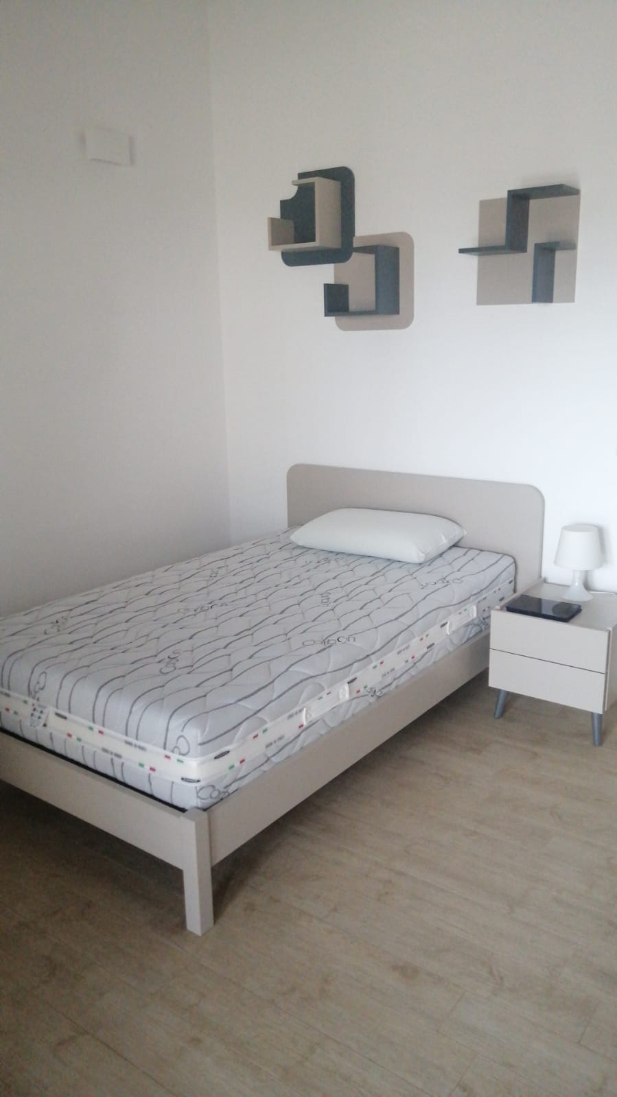 Camera da letto con letto singolo, comodino e mensole a muro contro una parete bianca.