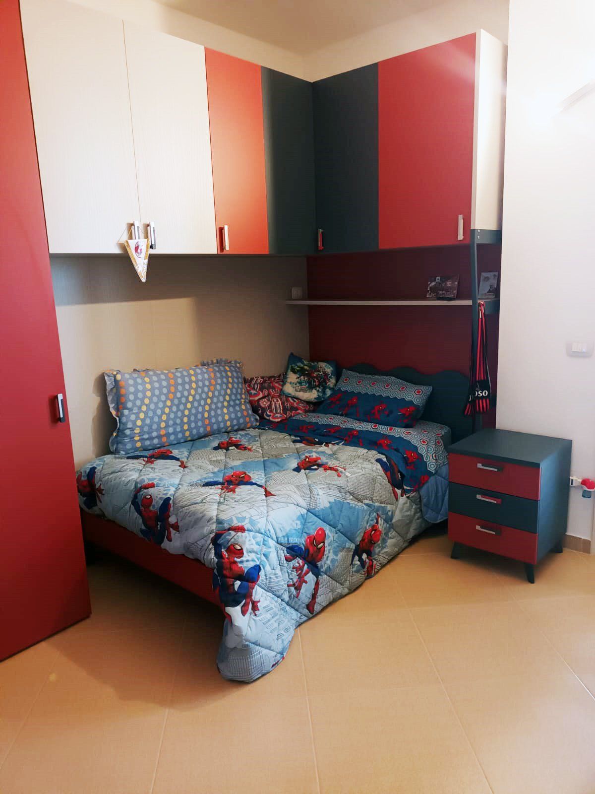 Camera da letto con letto a tema Spider-Man, dettagli rossi e blu, armadi a muro e comodino.