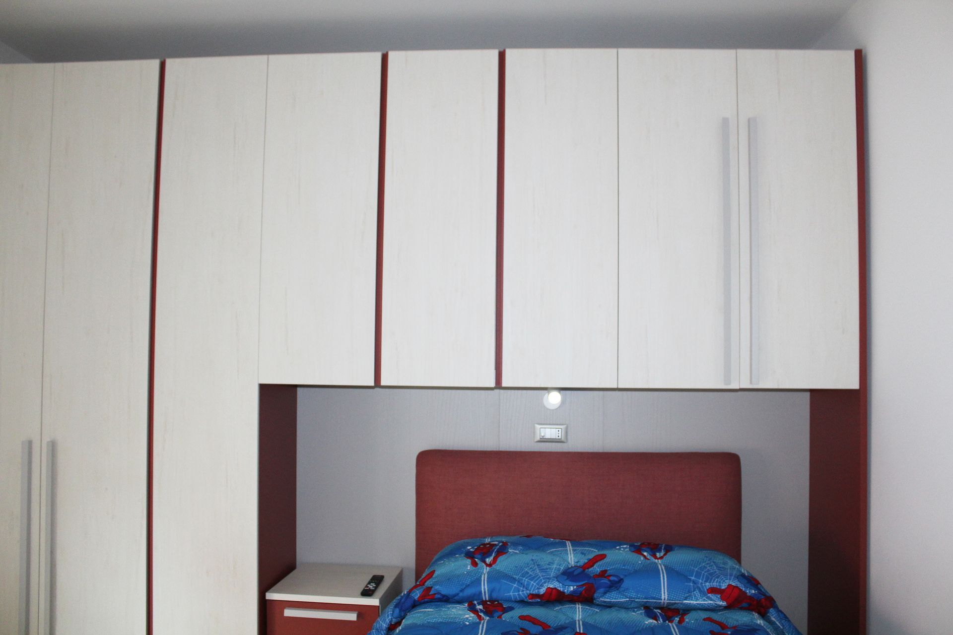 Camera da letto con armadi bianchi a muro e letto con struttura, comodino e finiture rossi.