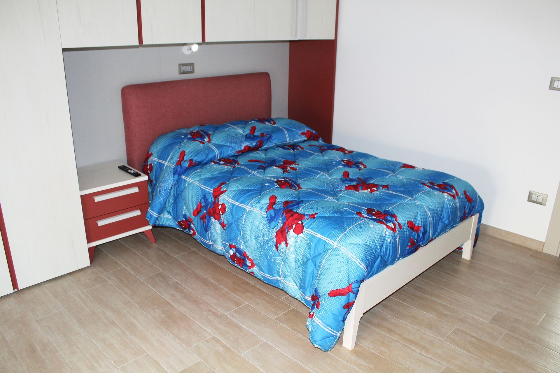 Camera da letto con piumone blu, struttura del letto bianca, testiera e comodino rossi e pensili.