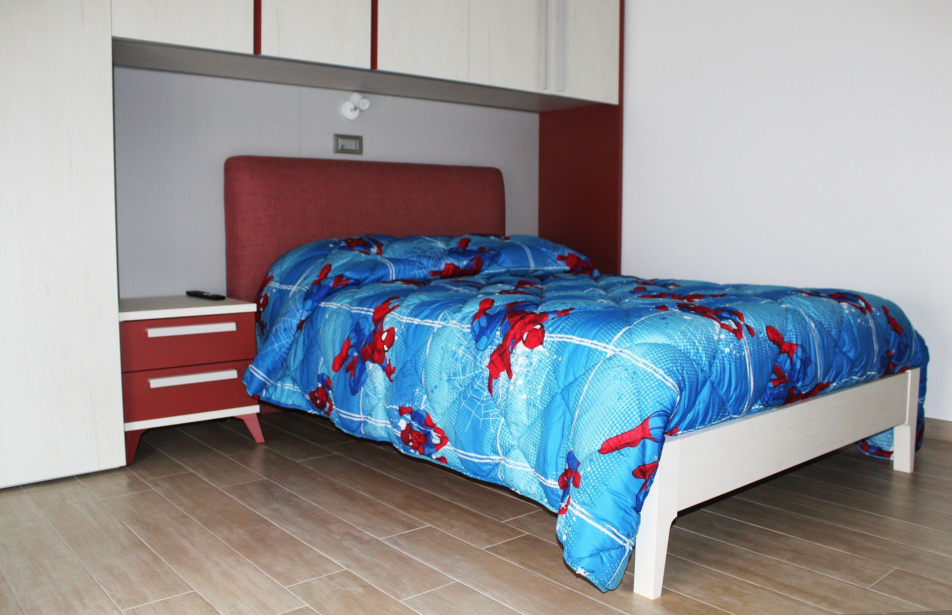 Camera da letto con letto, testiera rossa, piumone blu di Spider-Man, comodino rosso e mobili contenitori bianchi.