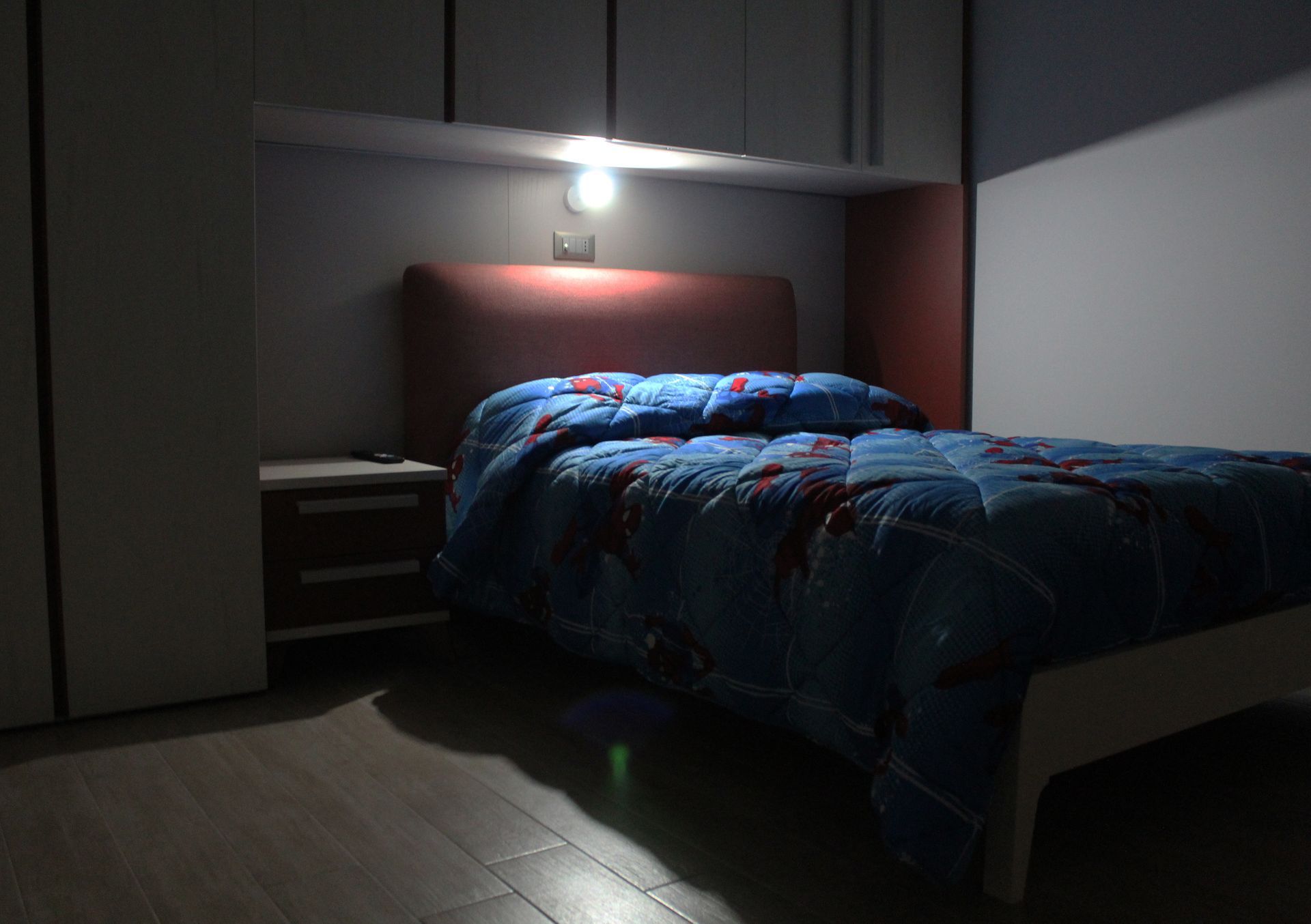 Camera da letto con letto, comodino e lampada da soffitto. La stanza è scarsamente illuminata, con lenzuola blu.