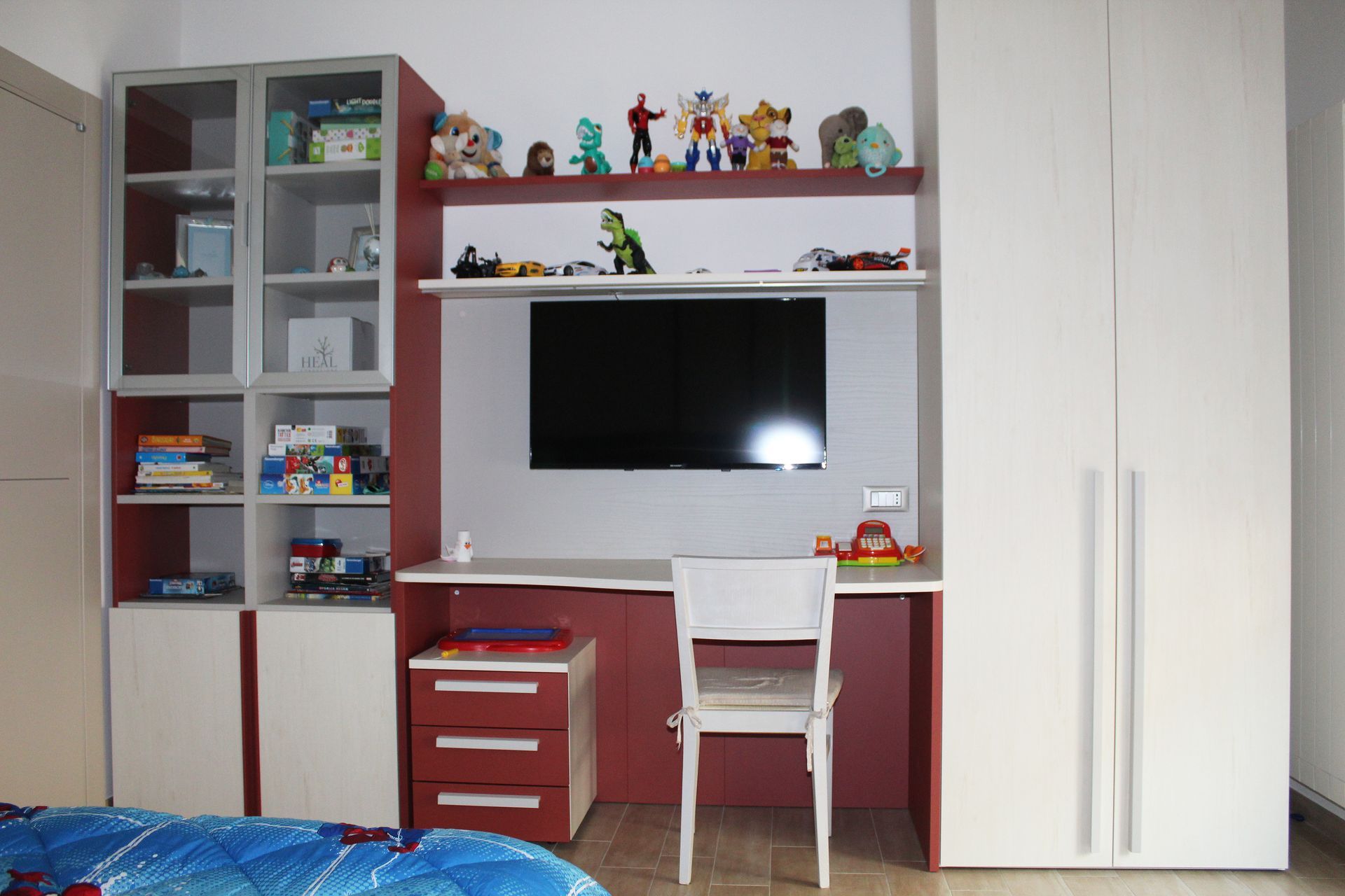 Una scrivania rossa e bianca per bambini con TV, circondata da armadietti bianchi e scaffali con giocattoli.
