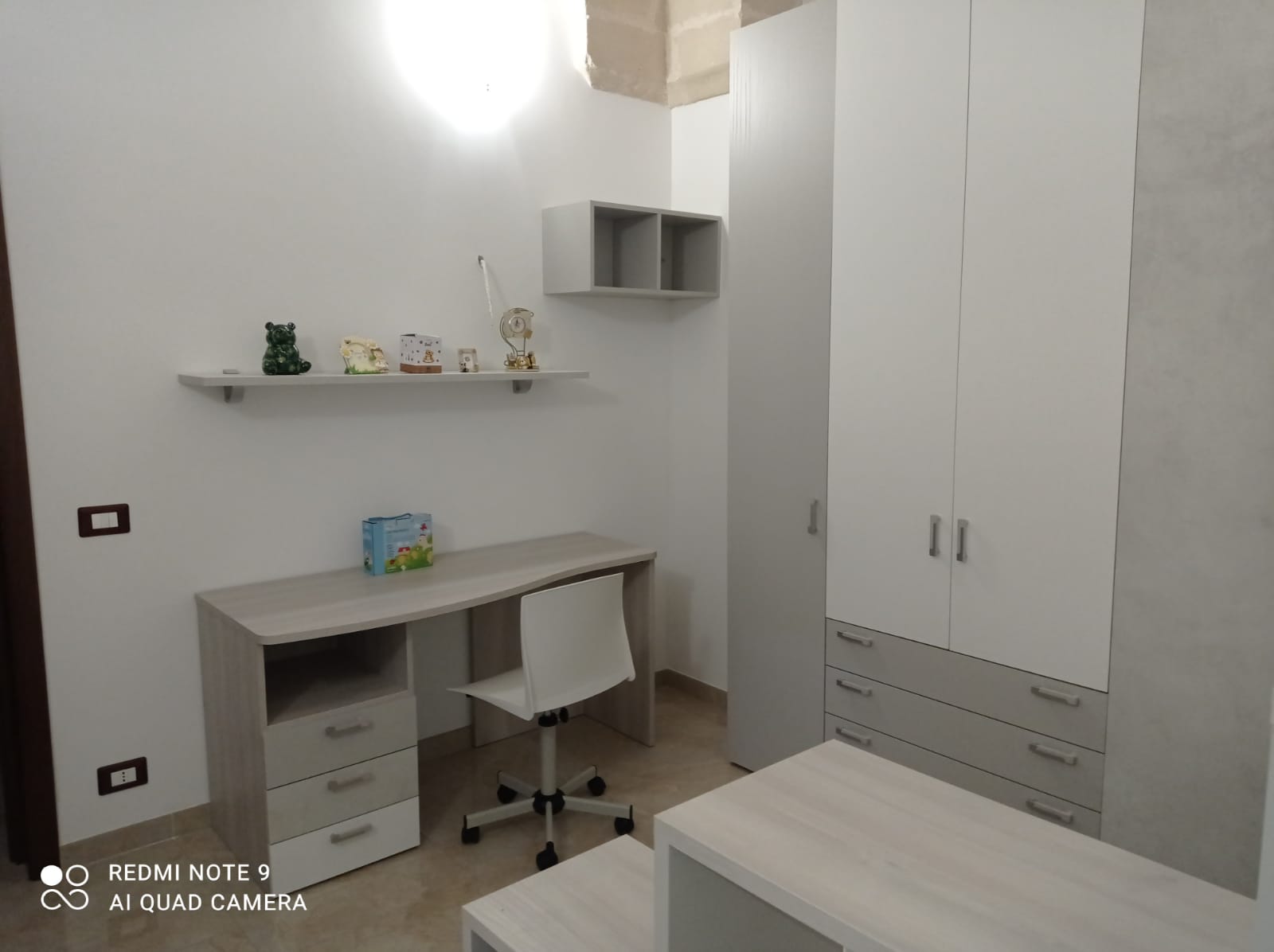Camera da letto con scrivania, sedia, armadio, scaffali e mobili bianchi e grigi.