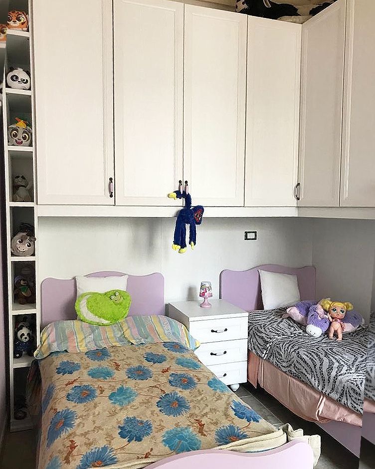 Cameretta per bambini con due letti, armadi bianchi e scaffali con animali di peluche.