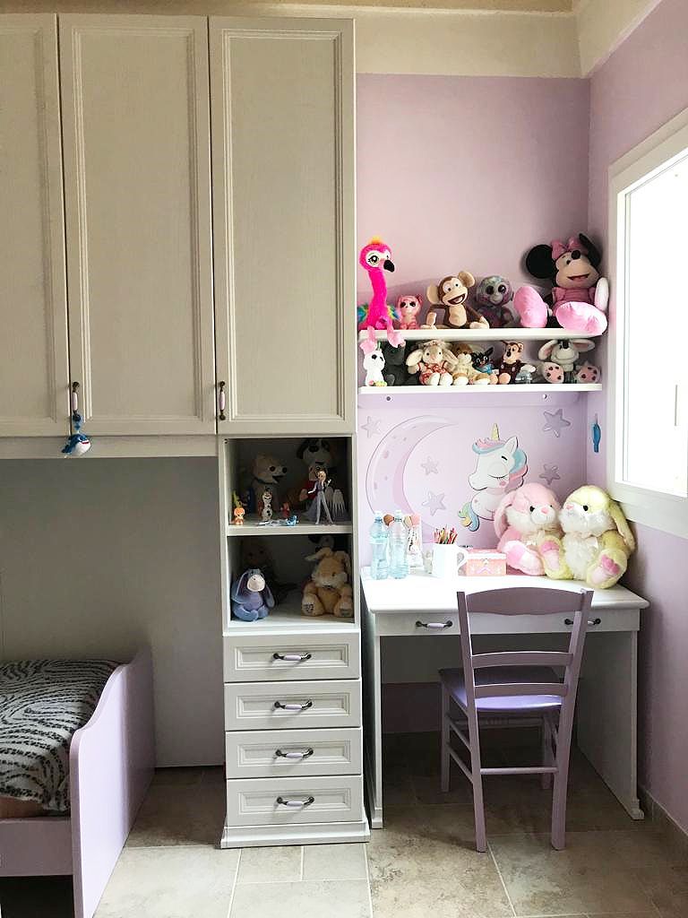 Cameretta per bambini con scrivania bianca, scaffali e armadio a muro. Colori pastello, peluche e un murale raffigurante un unicorno.