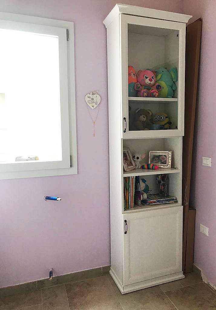 Mobiletto bianco con ante in vetro, dove sono esposti giocattoli e altri oggetti, accanto a una finestra su una parete color lavanda.