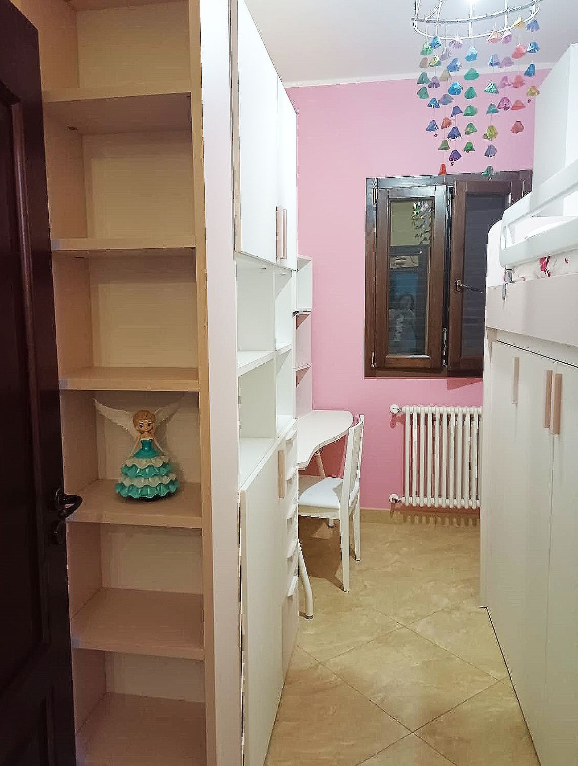 Una stanza con armadi bianchi, scaffali e una scrivania. Sono visibili pareti rosa, una finestra e un termosifone.