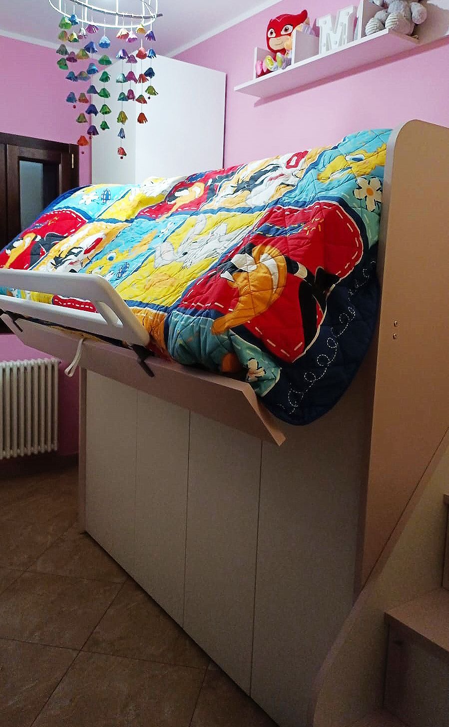 Nella cameretta di un bambino è presente un letto parzialmente rialzato con lenzuola colorate.