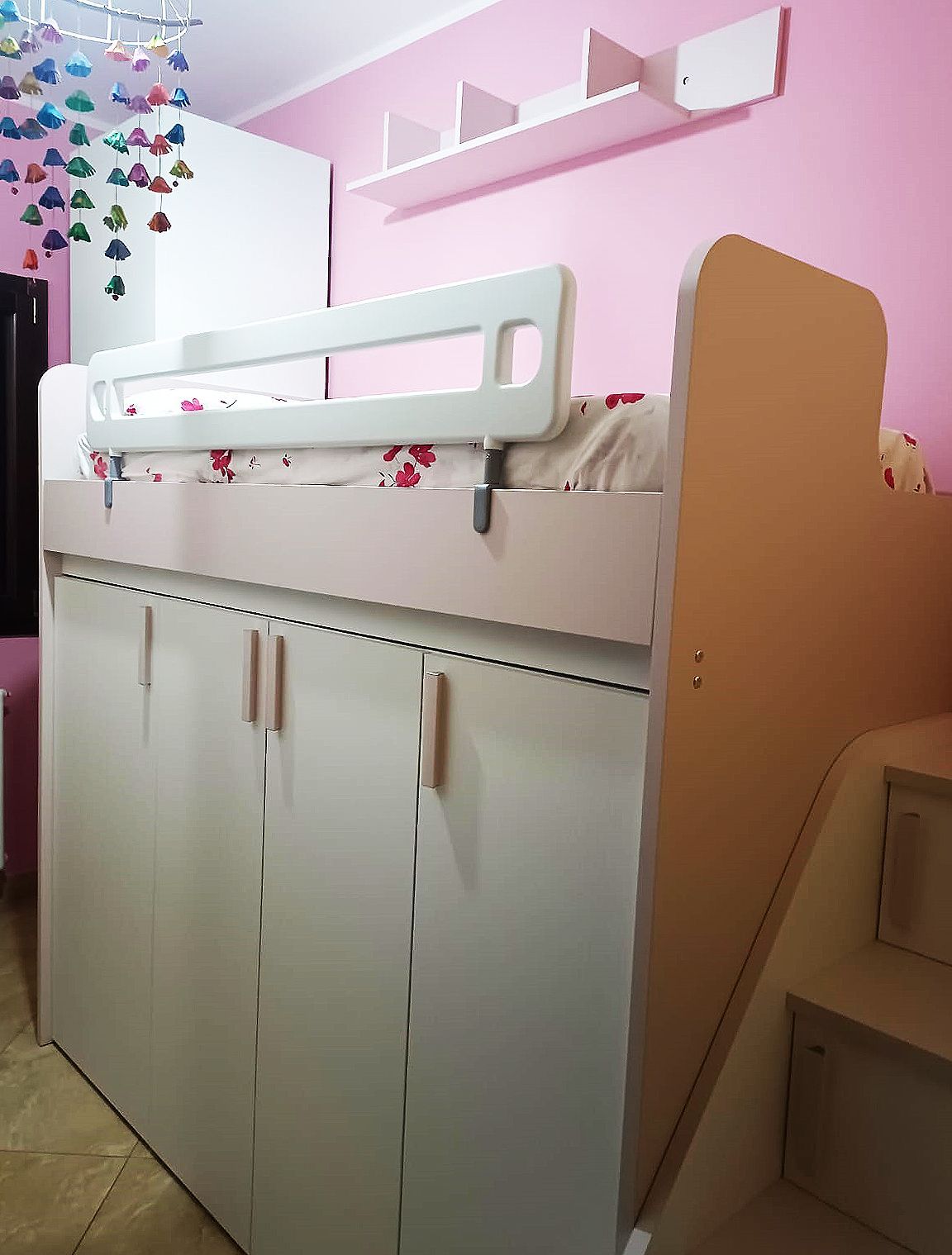 Letto a soppalco bianco con armadi a muro e spondina di sicurezza in una stanza rosa. Scale e scaffali in legno sono visibili.