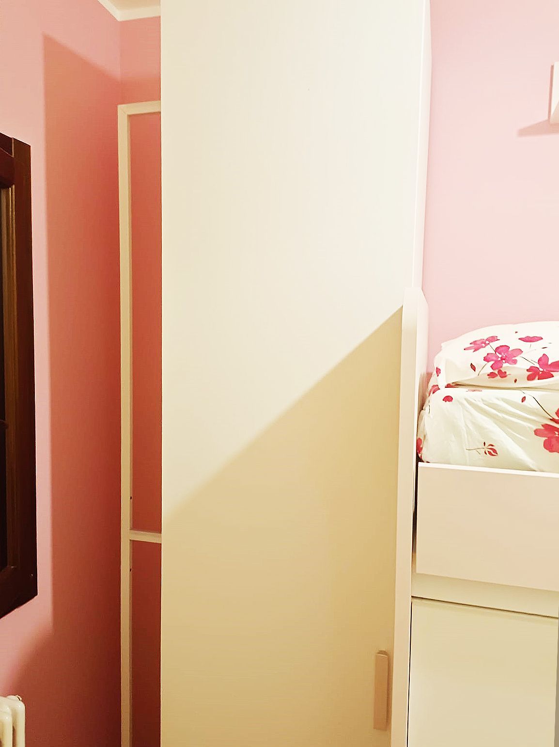Armadio bianco su pareti rosa; letto con biancheria floreale sulla destra.