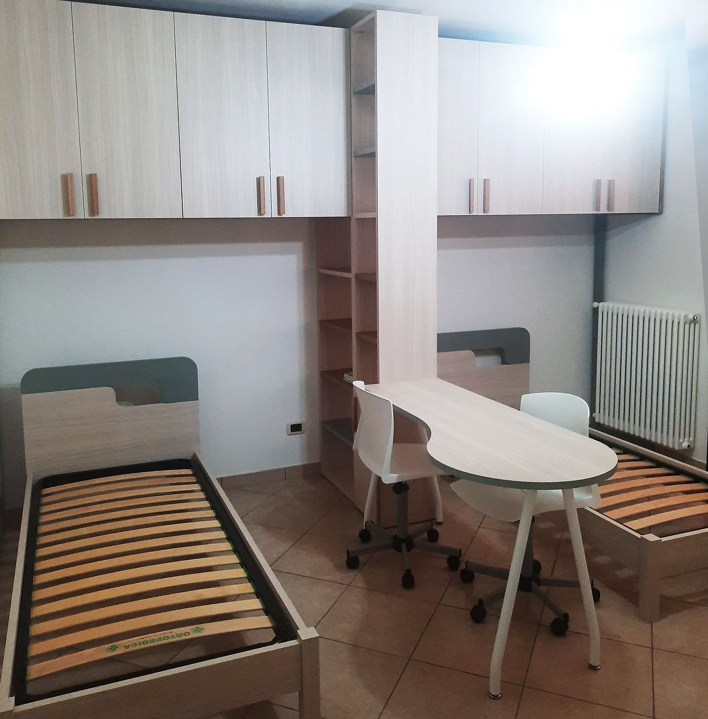Camera da letto con letto, scrivania, armadi e scaffali. Mobili in legno chiaro e pareti bianche.