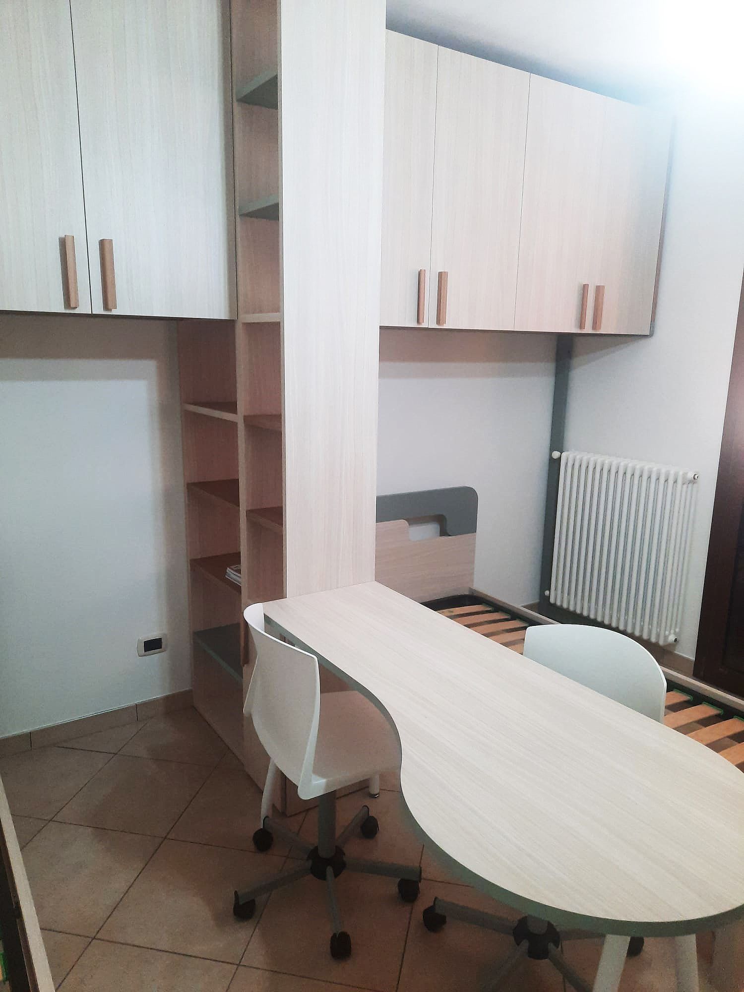 Una camera da letto arredata con scrivania, sedia, scaffali e armadi. Sono visibili mobili in legno chiaro e un termosifone.