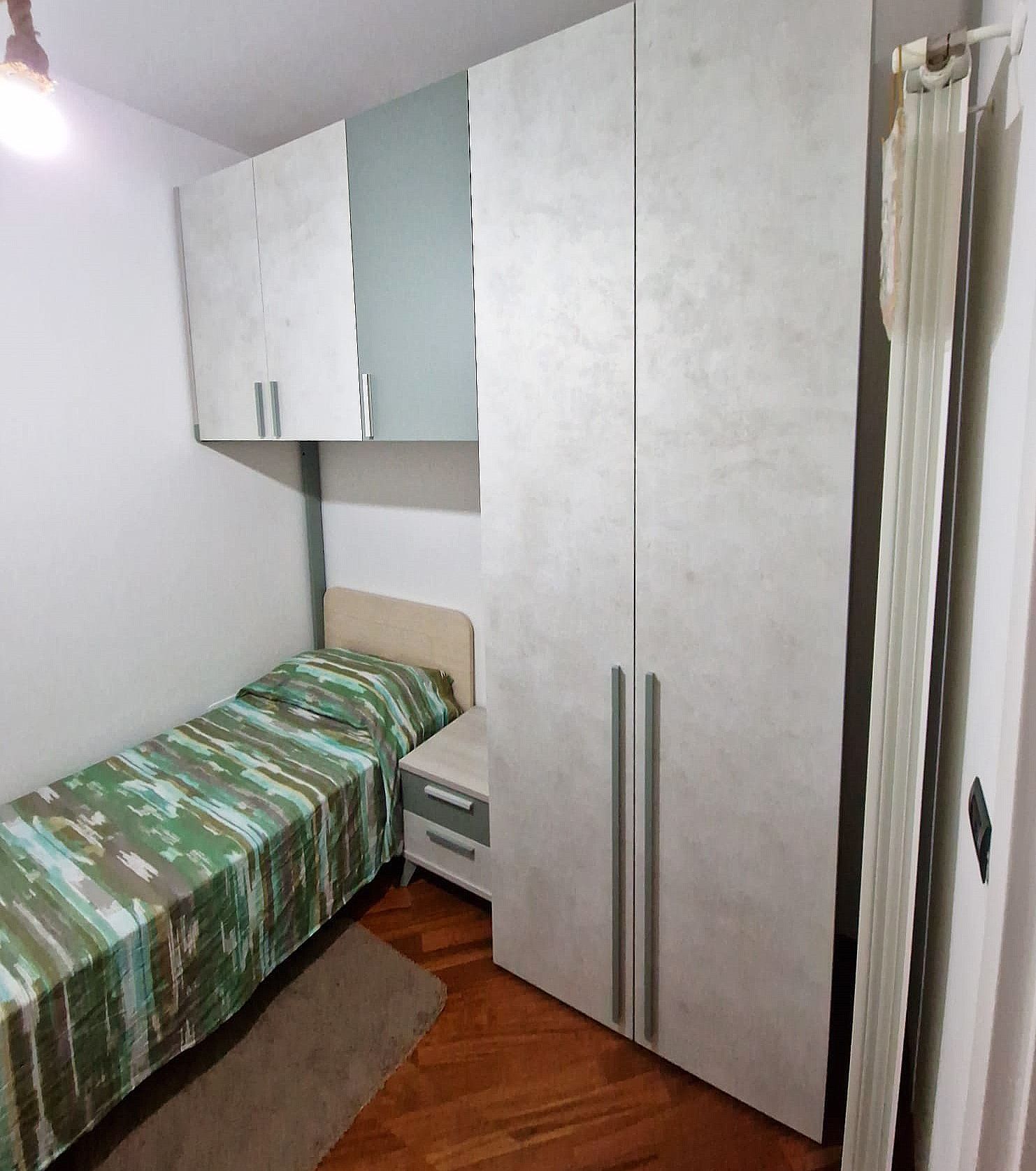 Piccola camera da letto con letto, armadio e pensili; biancheria da letto verde e bianca; pavimento in legno chiaro.