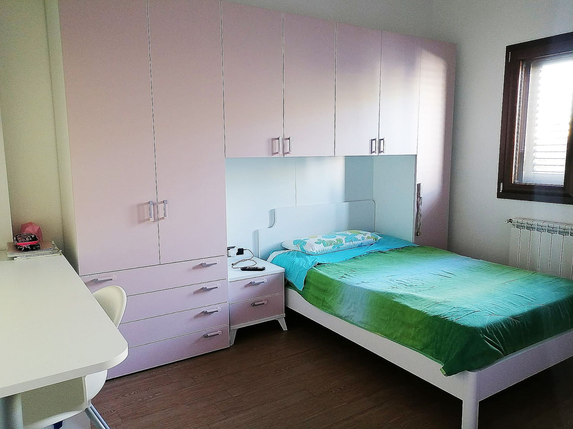 Una camera da letto rosa con letto, cassettiera, scrivania e armadio. Una piccola finestra lascia entrare la luce naturale.